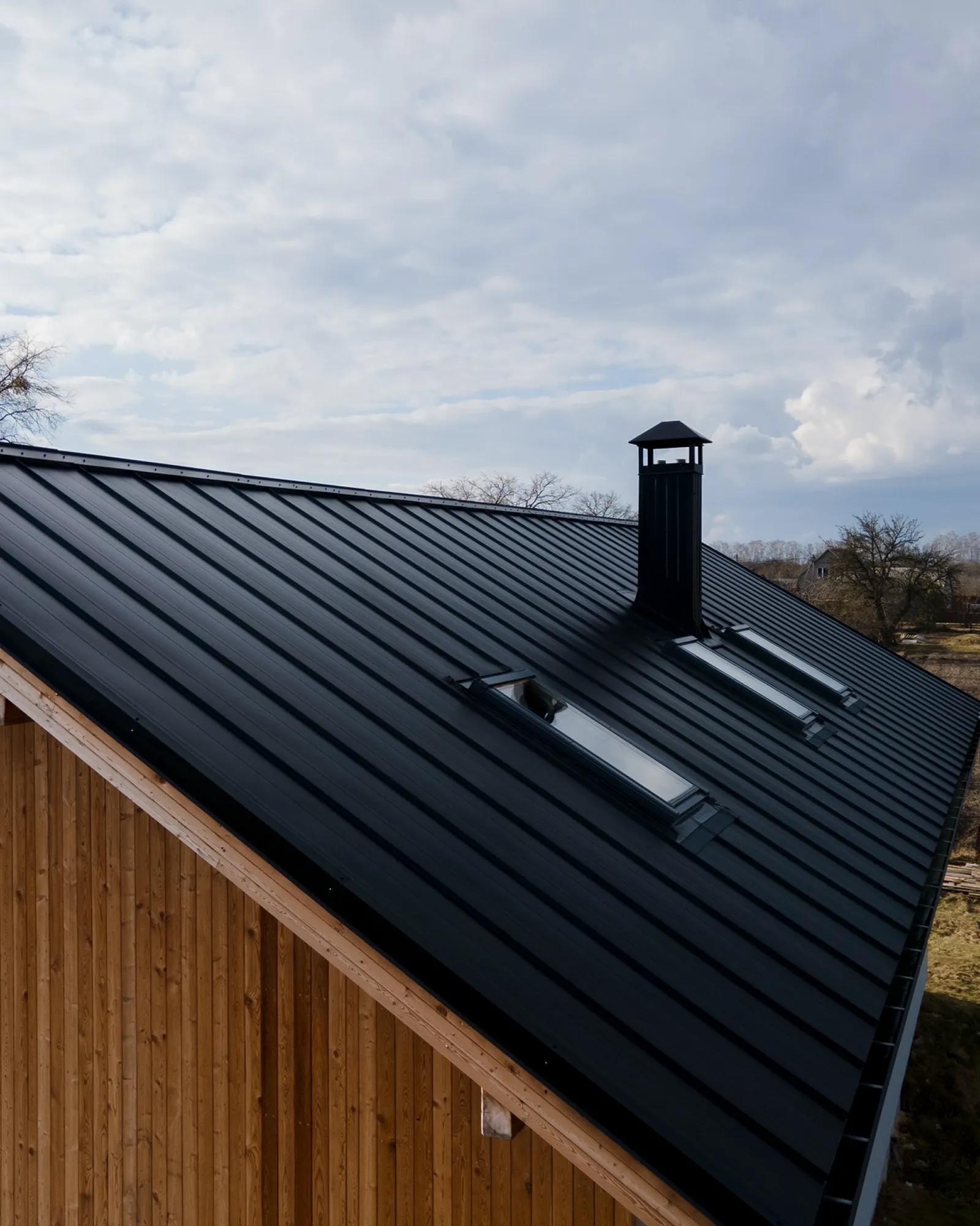 Modern black metal roof on a wooden house with two skylights and a chimney under a cloudy sky.